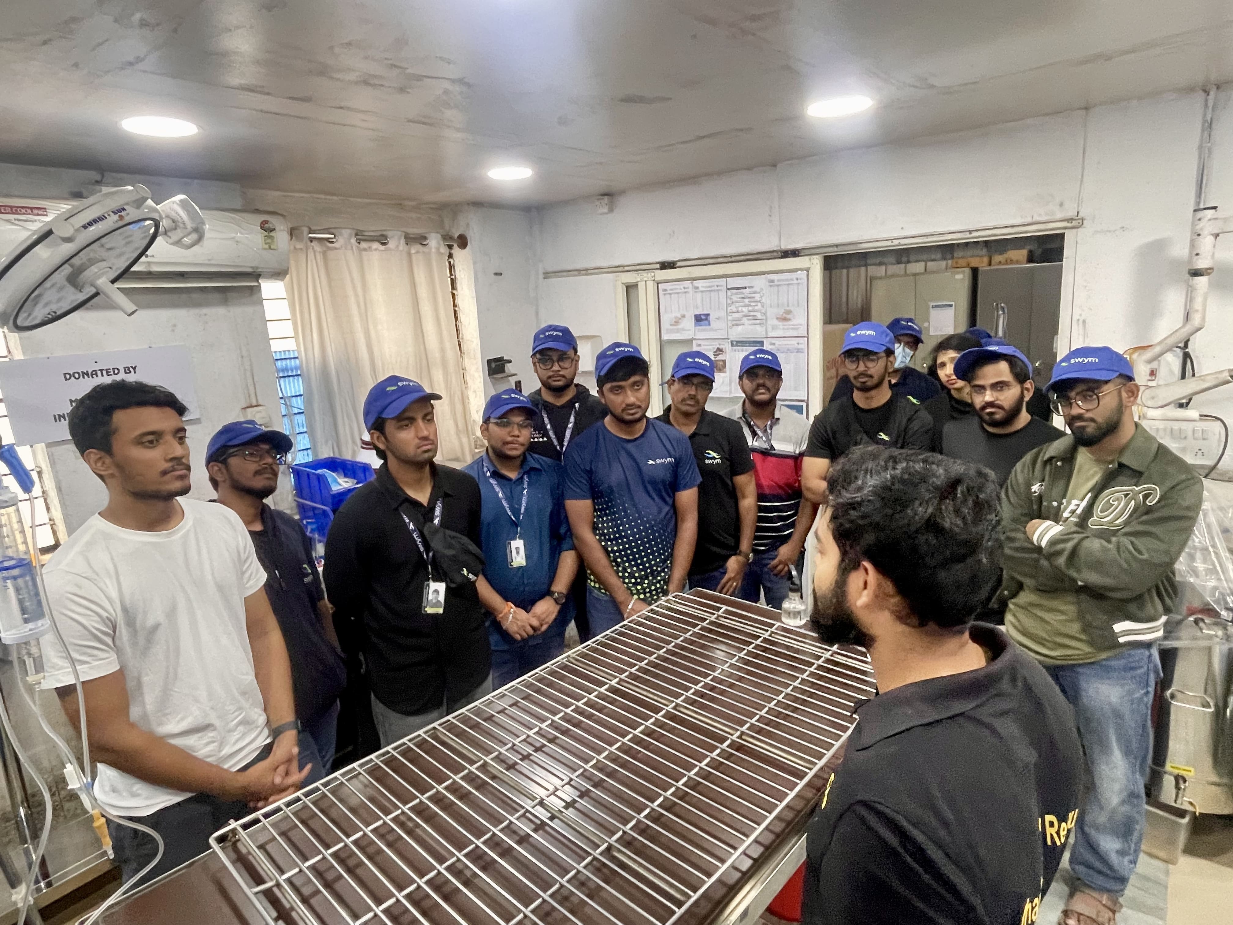 Group of young men standing around a medical examination table in a room, many wearing blue caps and badges, listening to a man facing them who wears a black shirt.