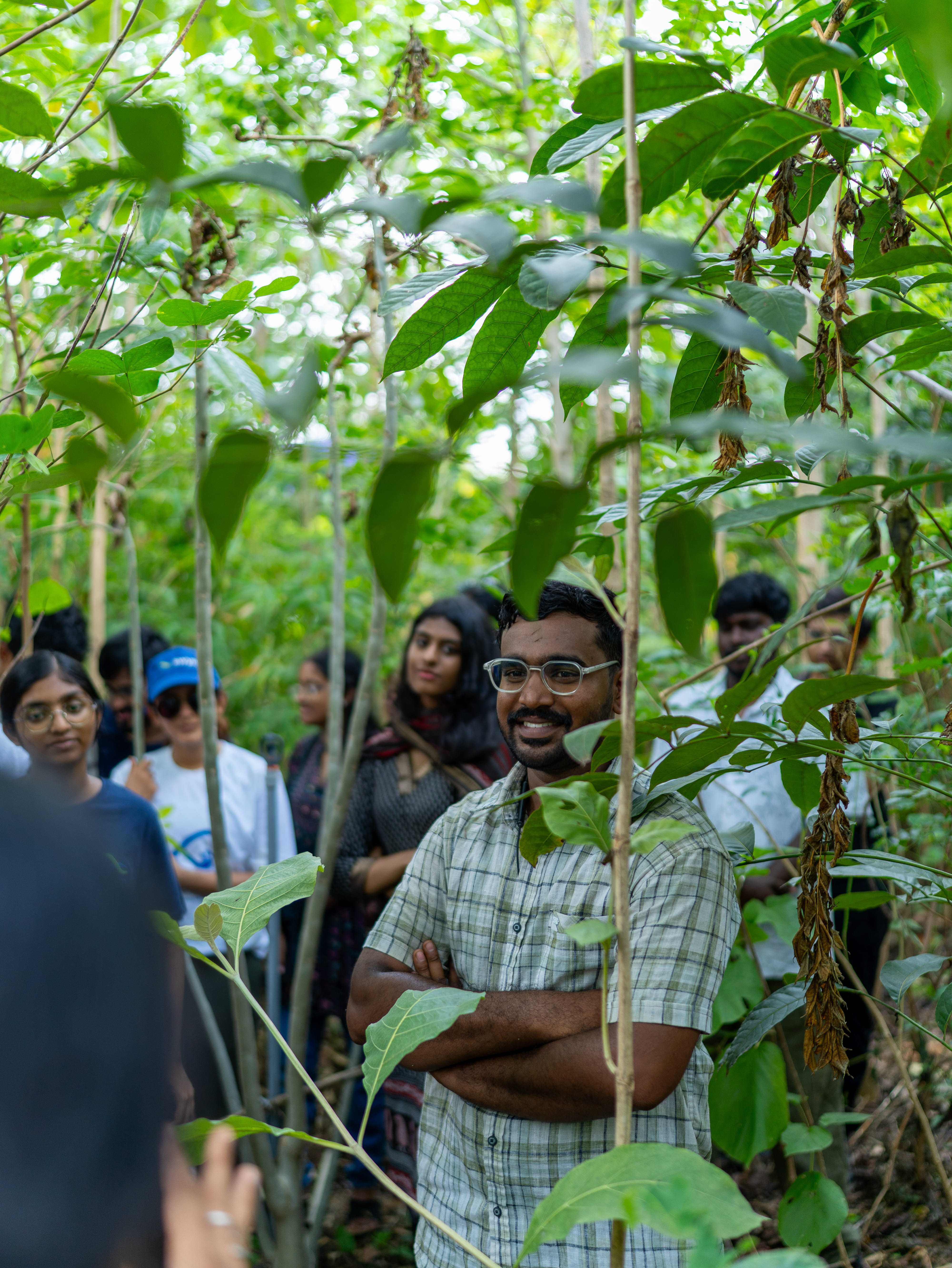 A man with glasses and a checkered shirt stands with arms crossed among dense green foliage while a group of people stand behind him in a wooded area.
