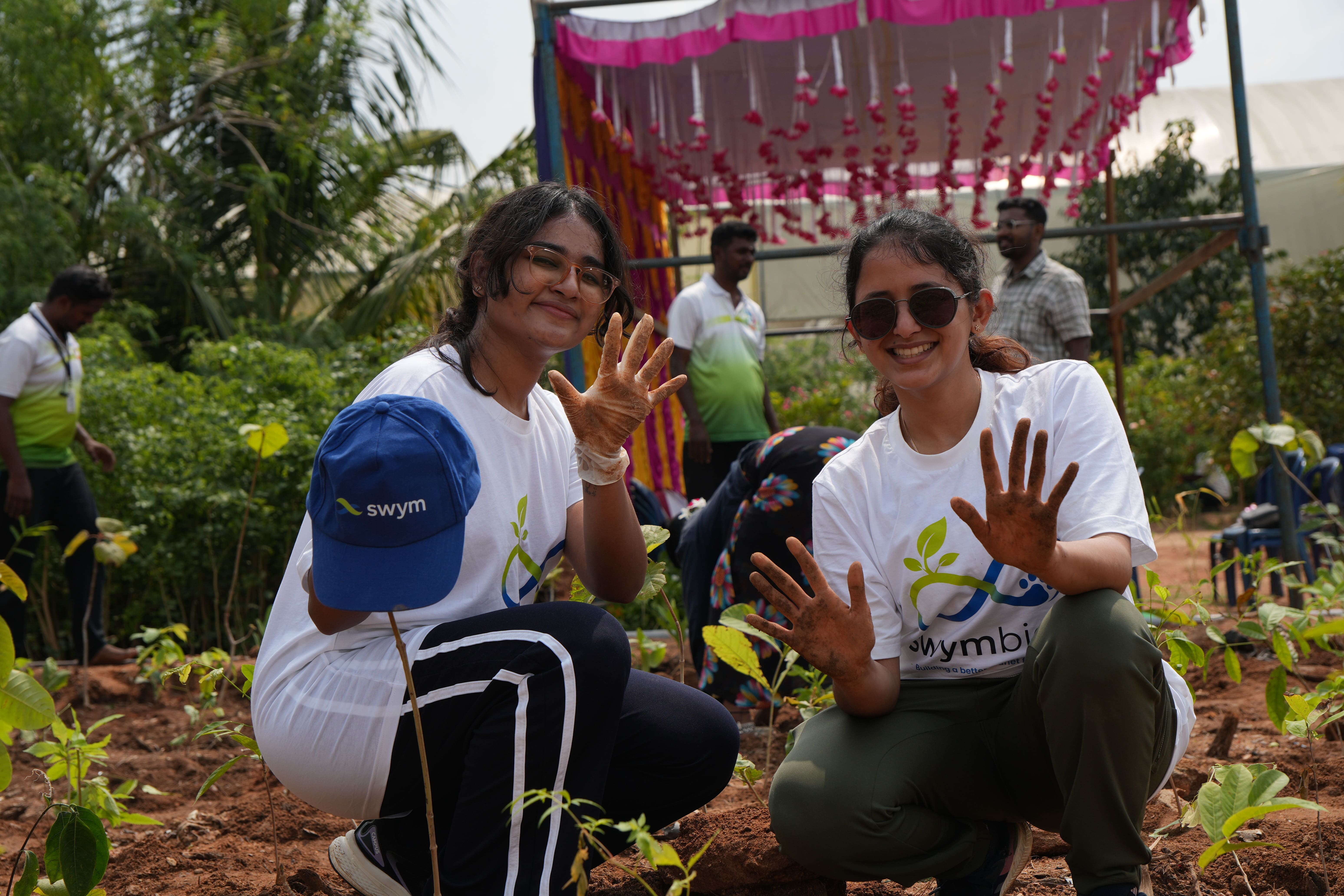 Two women wearing white Swym t-shirts and glasses smiling and showing their dirty hands while crouching in a garden with young plants, with other people and greenery in the background under a decorated canopy.