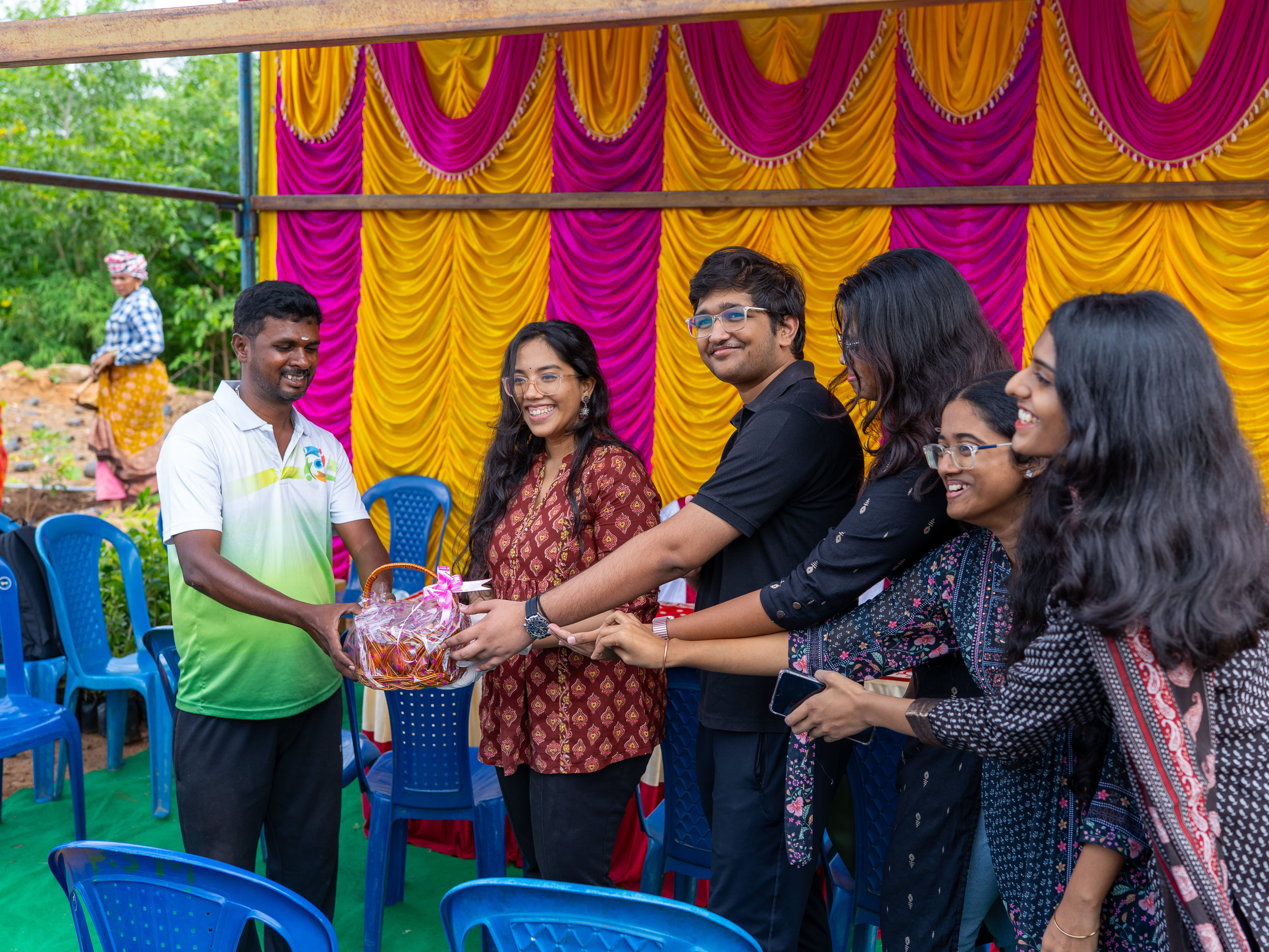 A group of five young people smiling and reaching to receive a gift basket from a man wearing a white and green shirt, with a vibrant pink and yellow draped backdrop behind them.