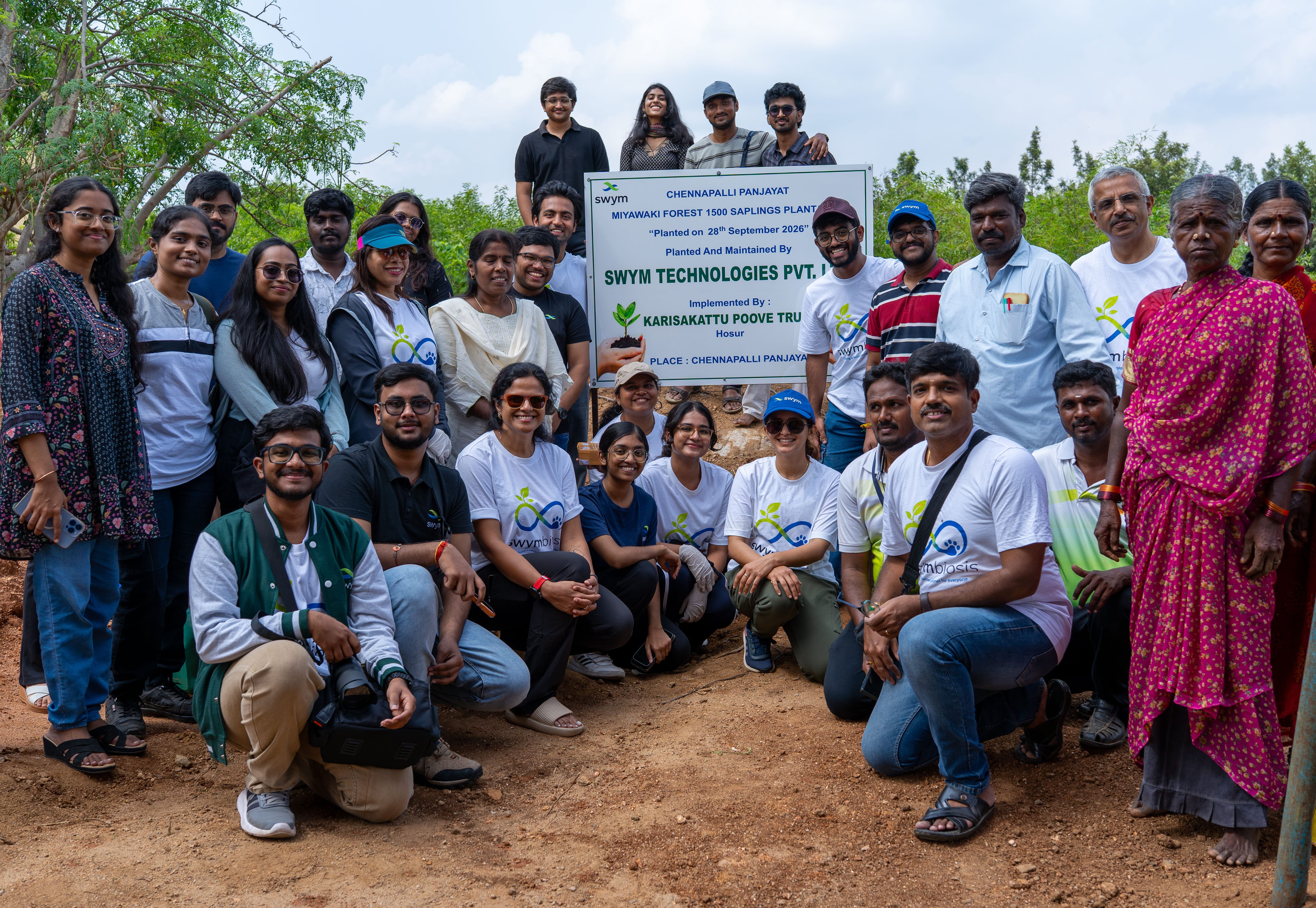 Group of people smiling and posing outdoors around a signboard that announces planting 1500 saplings in Miyawaki Forest, maintained by SWYM Technologies Pvt. Ltd. with Karisakattu Poove Trust at Chennapalli Panjayat on 28th September 2026.