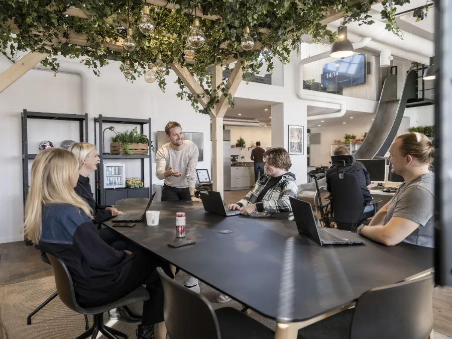 Ett team av kollegor sitter runt ett bord i ett modernt kontor och diskuterar arbete med laptops, med grönska och öppna ytor i bakgrunden.