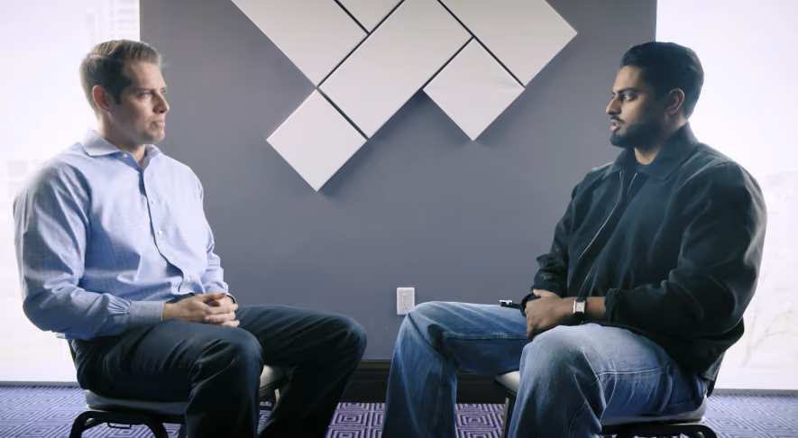 Two men seated facing each other in a modern office with geometric wall art behind them.