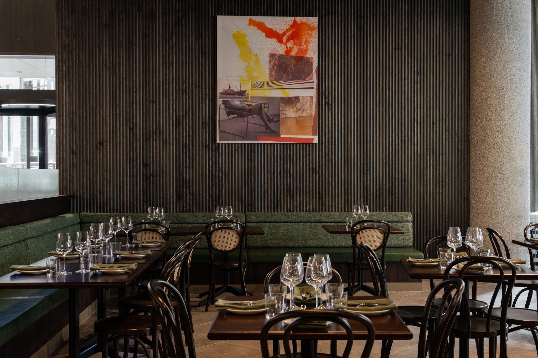 Modern restaurant interior with dark wooden tables, green cushioned bench seating, black bentwood chairs, and an abstract colorful painting on a dark corrugated wall.