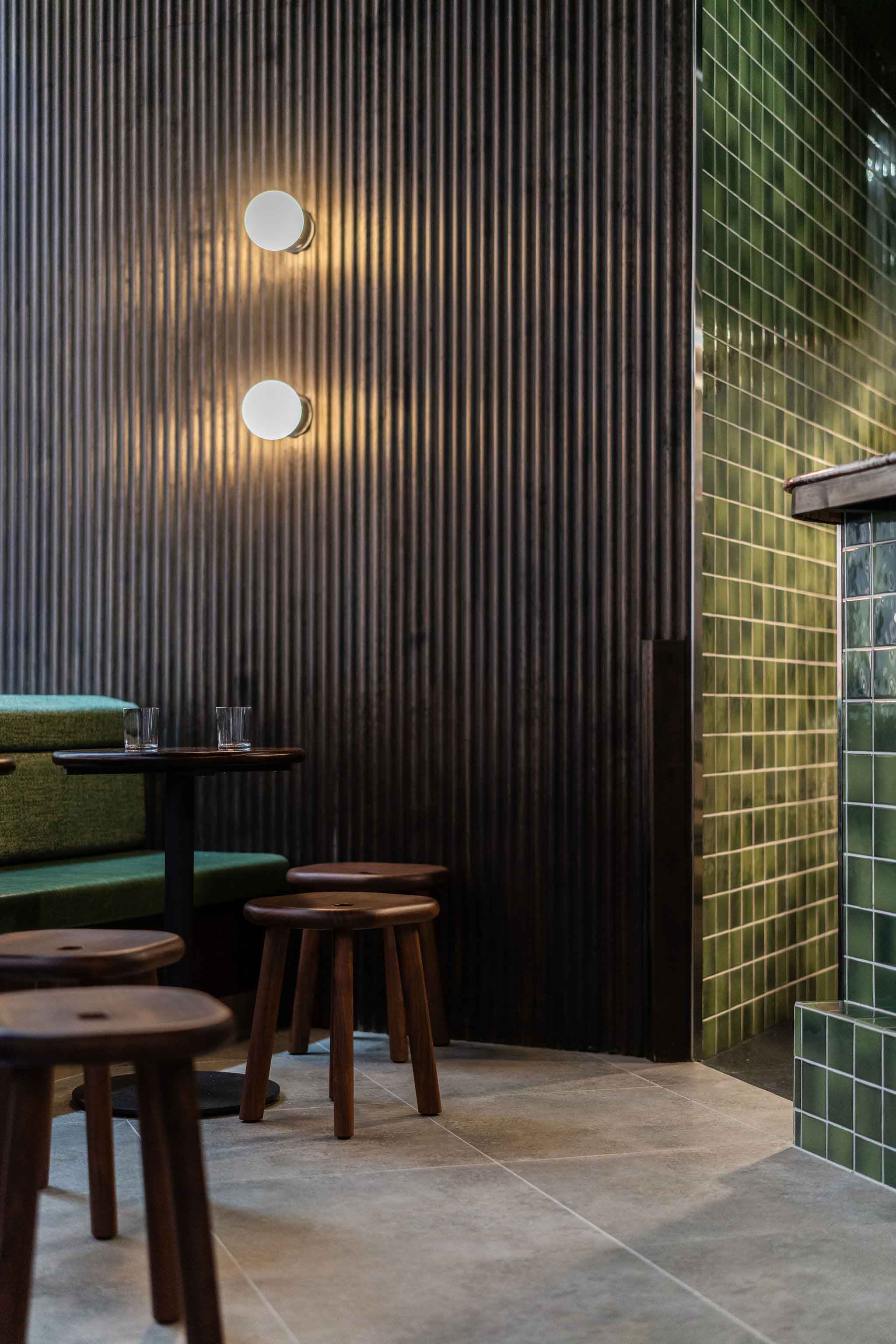 Modern café corner with dark corrugated wall, green tiled counter, wooden stools, and a small tables with glasses.