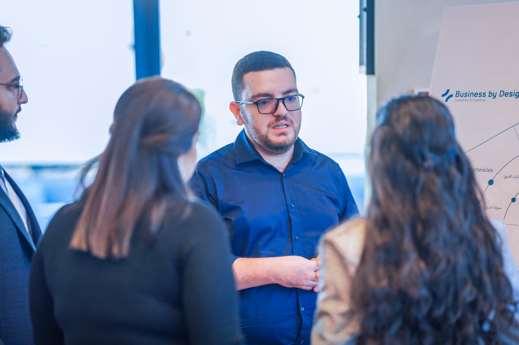 A man in glasses speaks to three people near a business presentation board.