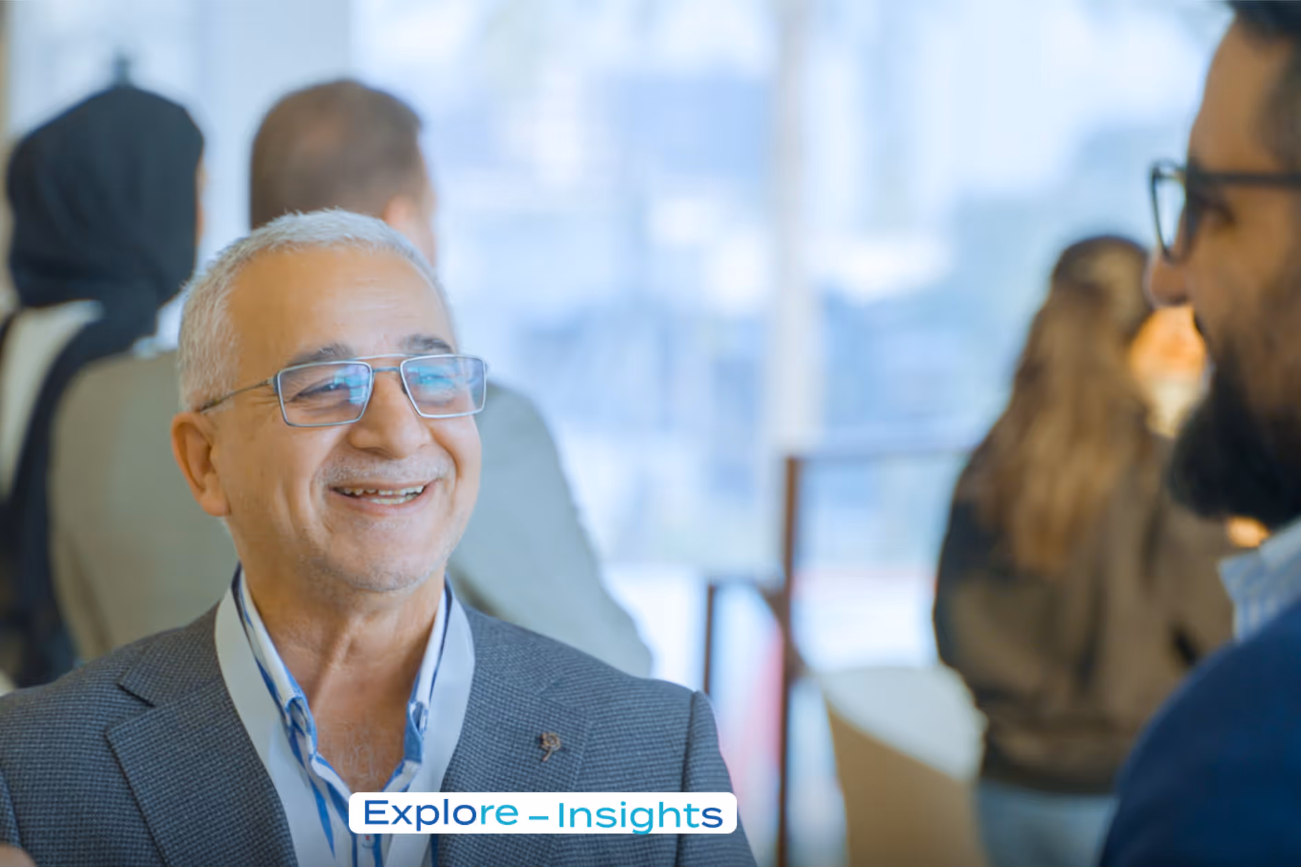 An older man with glasses smiles while talking to another man during a casual event.