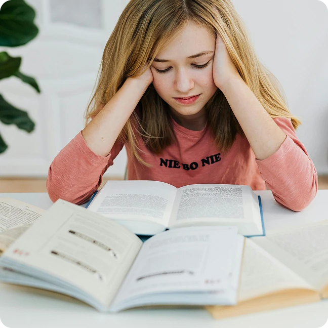 Jeune fille concentrée lisant un livre avec plusieurs livres ouverts devant elle sur une table.