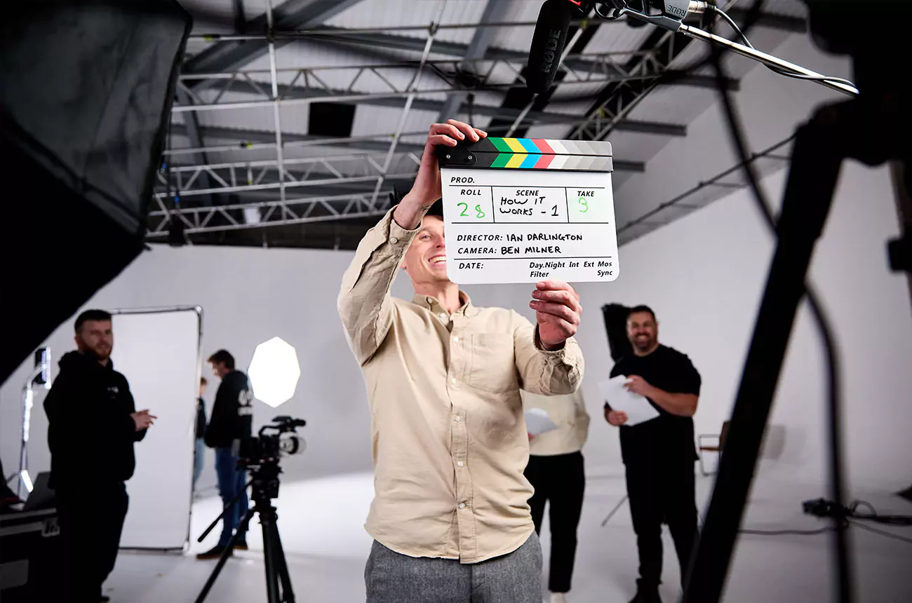 Sean Waring, Studio Director at GNG Studios, holding a clapperboard on set with crew and lighting setup behind him.