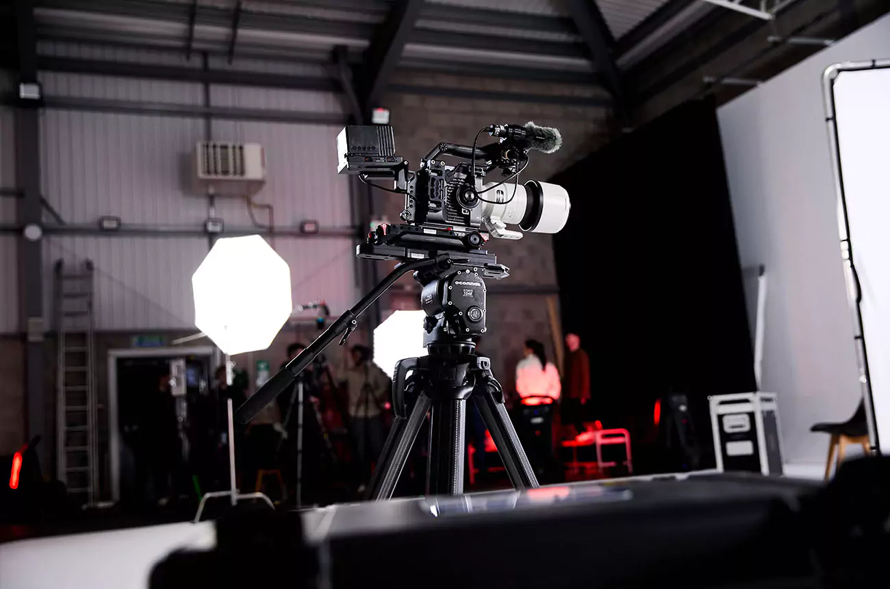 Close-up of professional cinema camera on tripod at GNG Studios, with lights and crew preparing for a video shoot in the background.