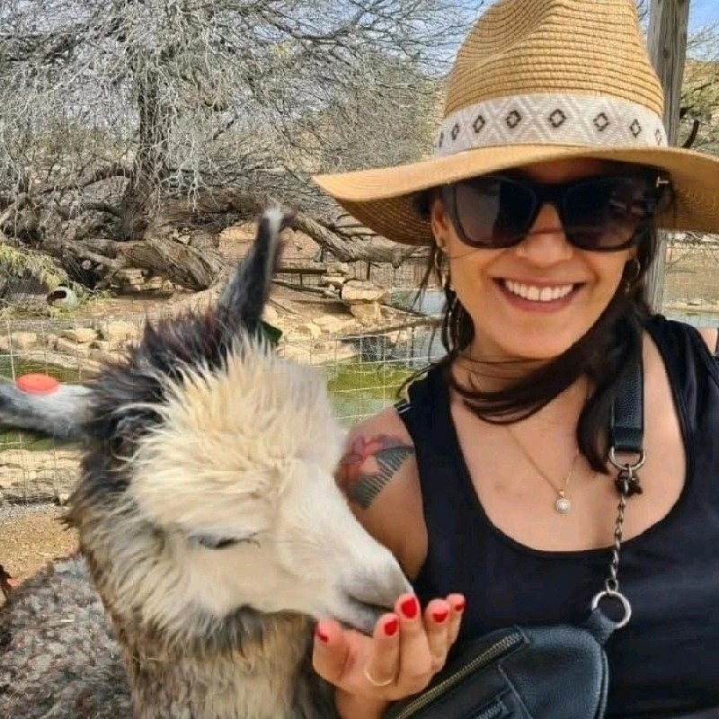 Smiling woman wearing sunglasses and a wide-brimmed hat feeding a white and gray llama with red-painted nails.