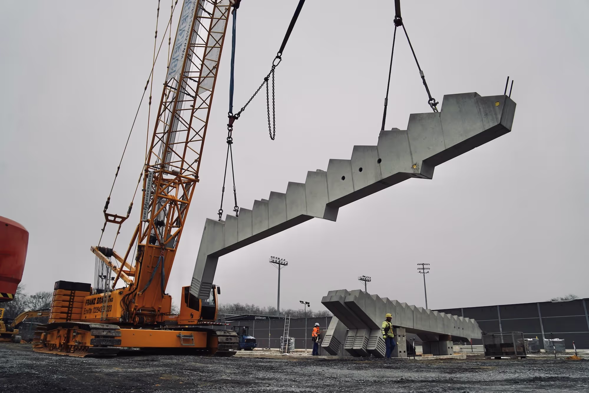 Ein großer Kran hebt ein massives Fertigteil-Betontreppenelement auf einer Baustelle, während Bauarbeiter in Schutzkleidung die Montage einweisen.