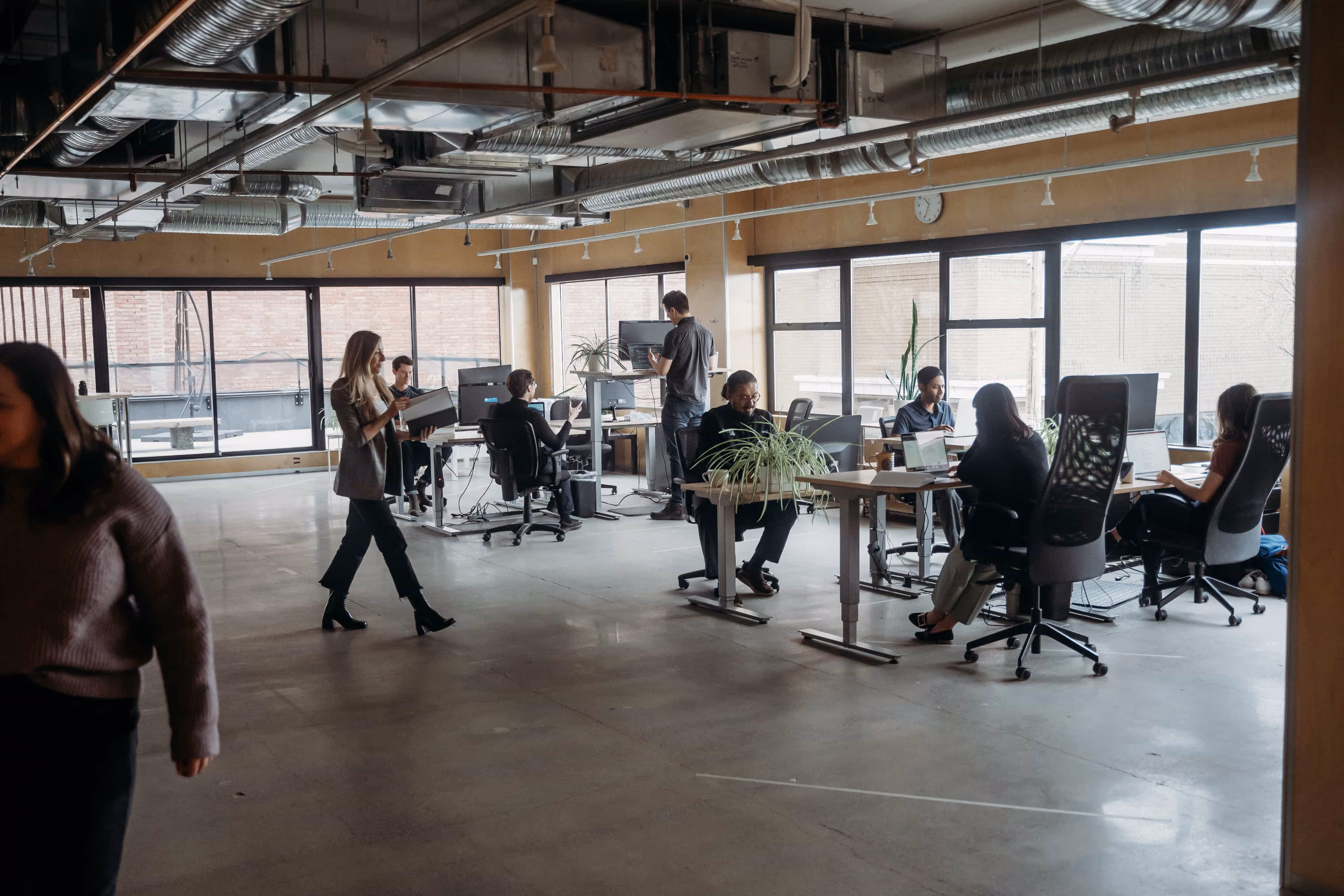 Open office space with people working at desks and a woman walking while holding a folder.