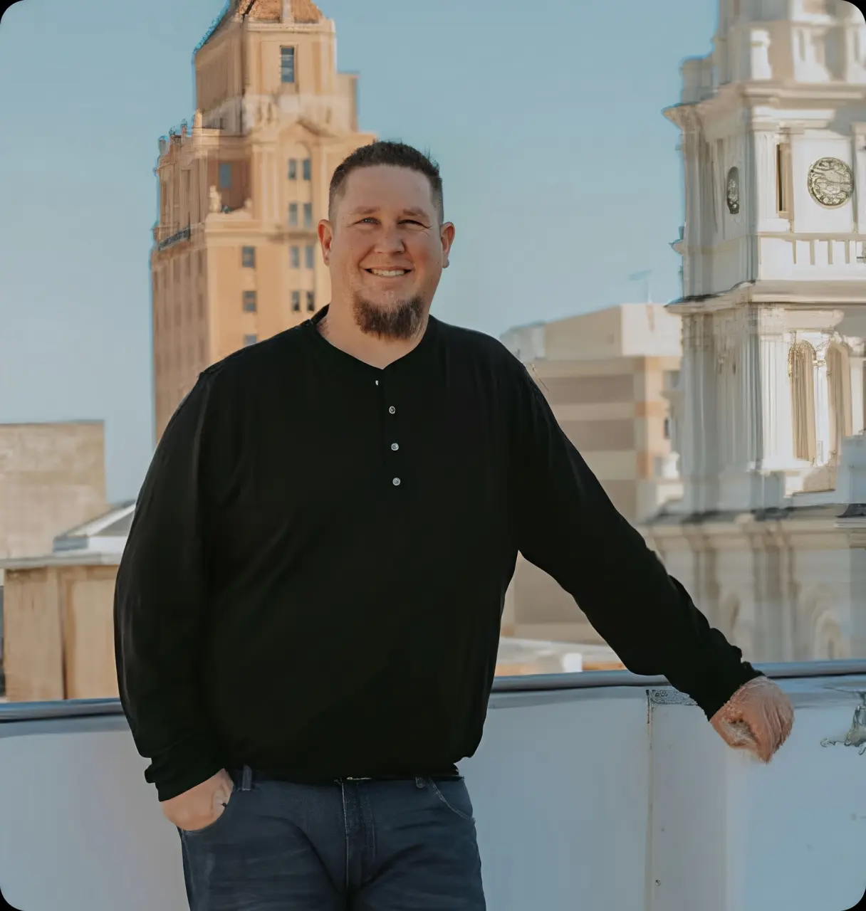 A bearded man in a black long-sleeve shirt and jeans smiles and leans against a white railing on a rooftop with tall city buildings in the background.