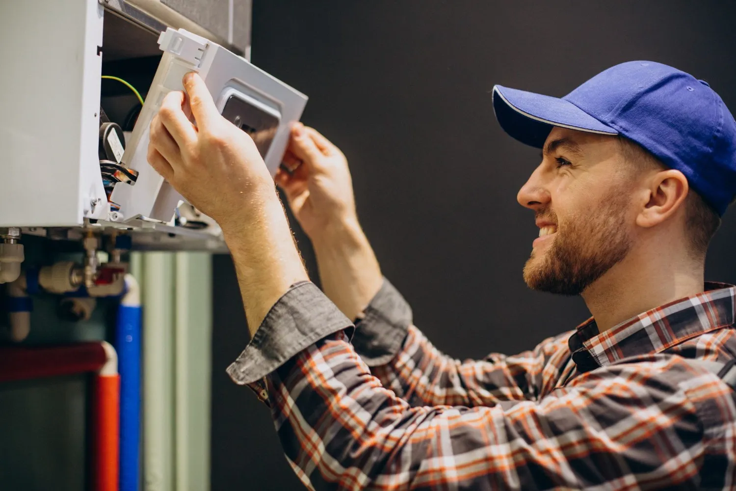  A man wearing a blue hat repairs a water heater in a home setting.