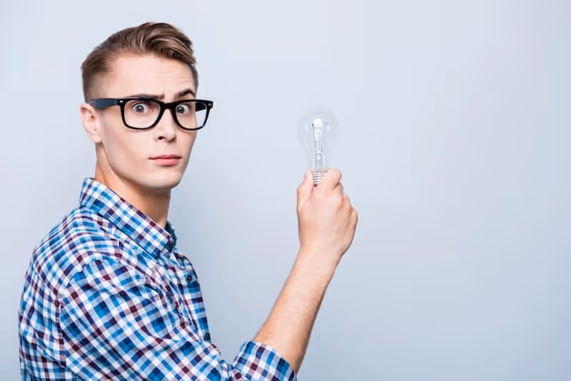 A man with glasses is holding a light bulb, looking thoughtfully at it.