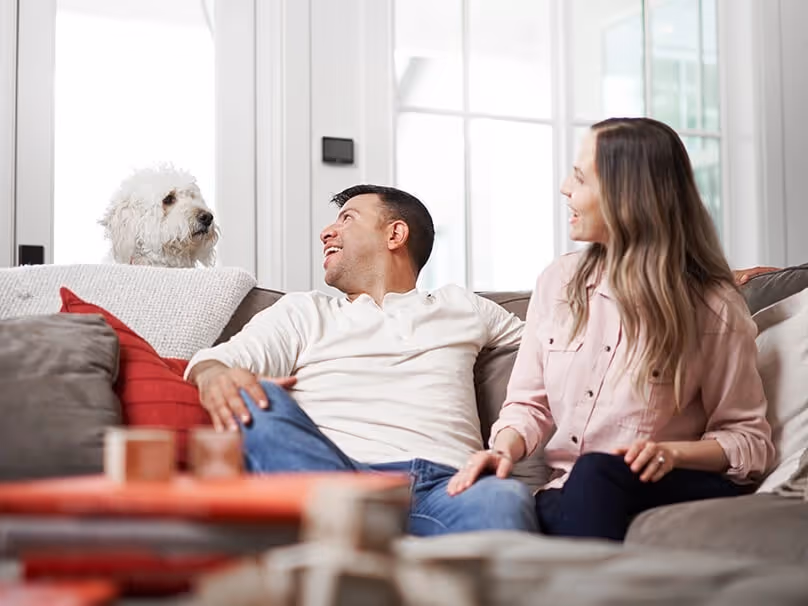 A couple sits on a couch, smiling at each other and a white dog.