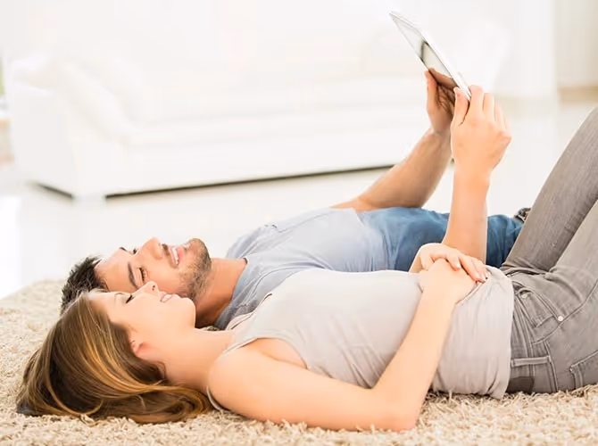 A man and woman lying on the floor, both focused on a tablet in front of them.
