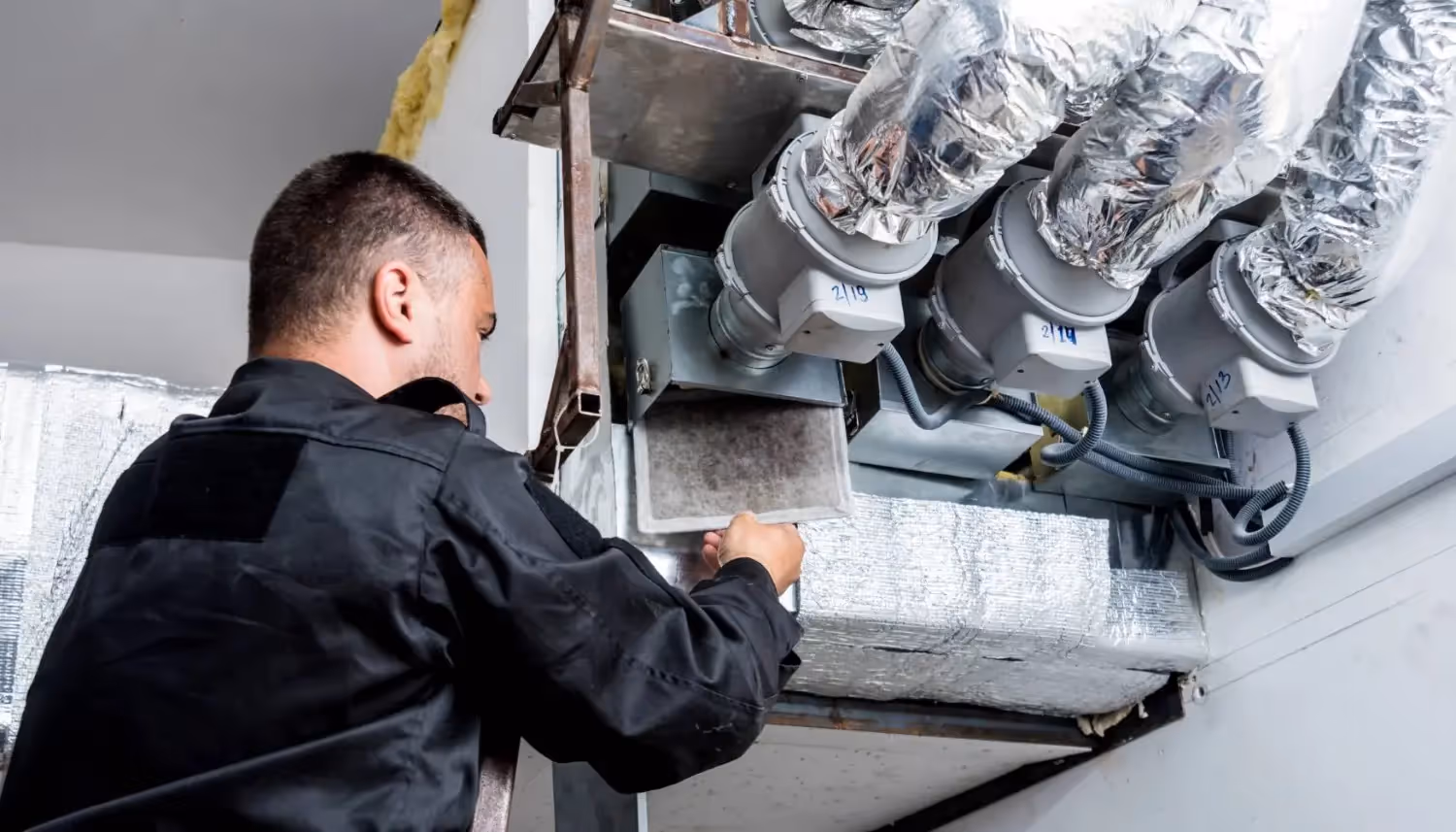 A technician in a black uniform inspects a ventilation system with insulated ducts and electrical components.