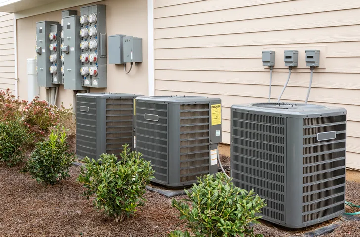 Three outdoor air conditioning units are lined up next to a building with siding, near several electricity meters.