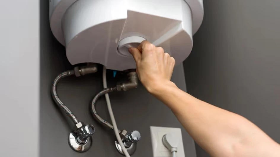 A hand adjusts the control on a white water heater mounted on the wall, with pipes and valves visible below.