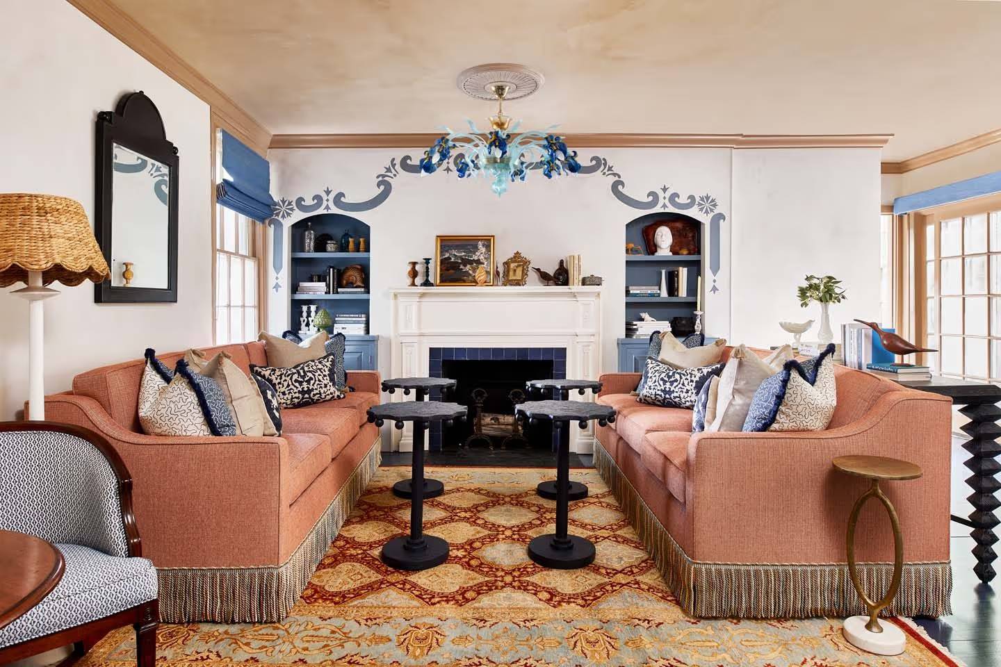 A traditional living room with two pink sofas, four black side tables, a fireplace, bookshelves, and blue decorative accents.