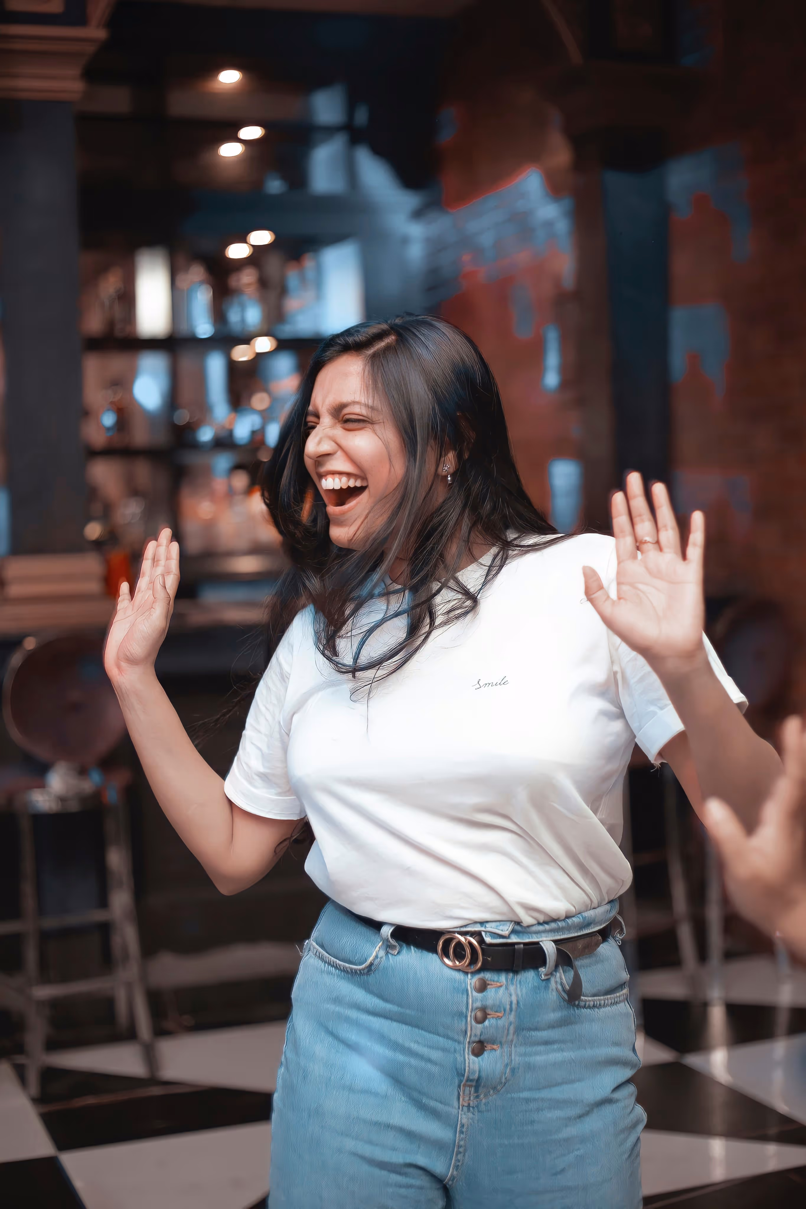 Femme souriante avec les bras levés, portant un t-shirt blanc et un jean bleu dans un intérieur.