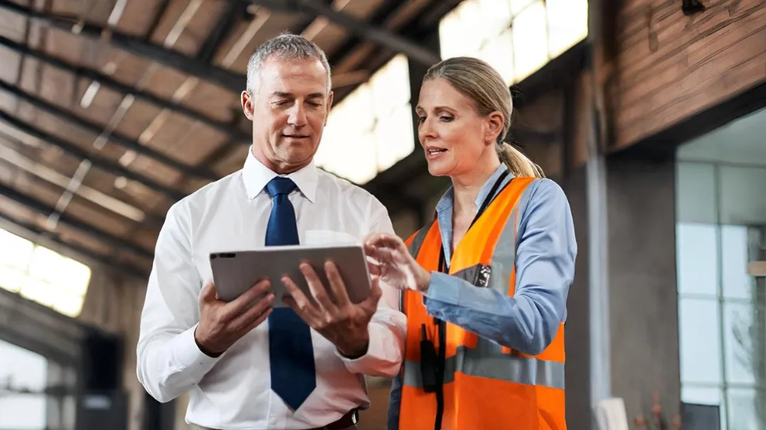 Zwei Personen, ein Mann im weißen Hemd mit blauer Krawatte und eine Frau mit oranger Warnweste, betrachten zusammen ein Tablet in einer industriellen Halle.