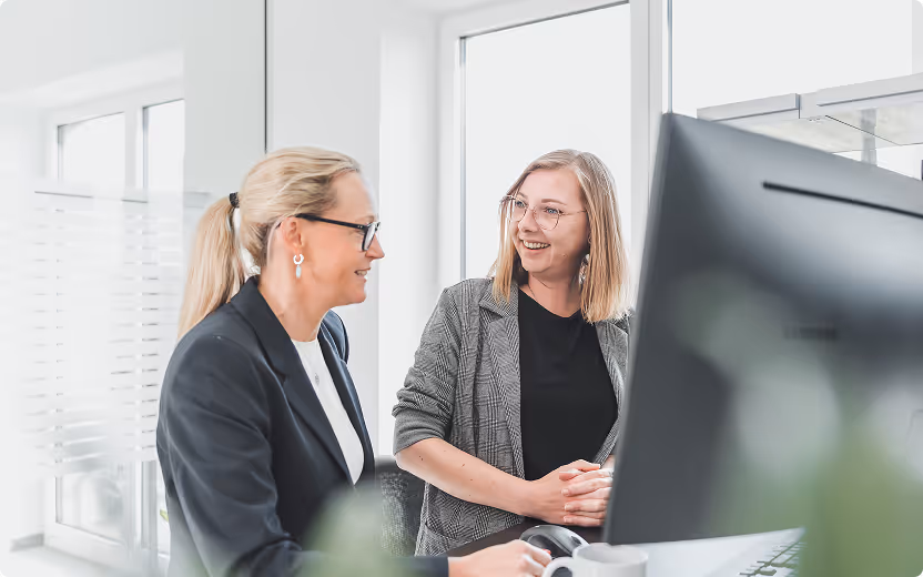 Zwei lächelnde Frauen im Büro unterhalten sich vor einem Computerbildschirm.
