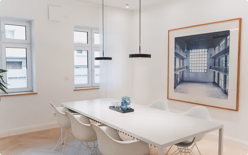 Moderner, heller Konferenzraum mit weißem Tisch, sechs weißen Stühlen und großem Bild an der Wand.