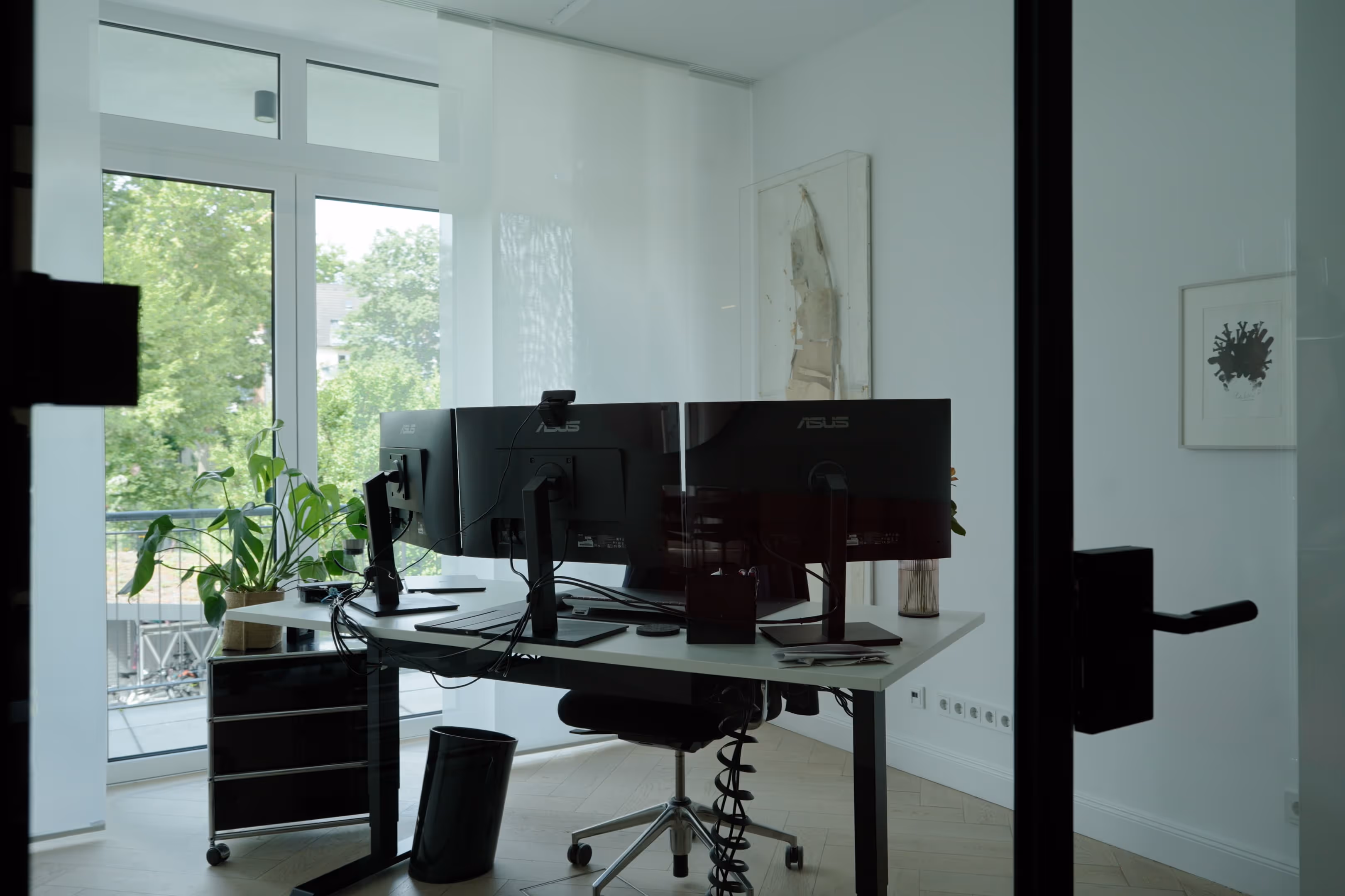 Modernes Büro mit zwei ASUS-Monitoren auf einem weißen Schreibtisch vor großen Fenstern mit Blick auf grüne Bäume.