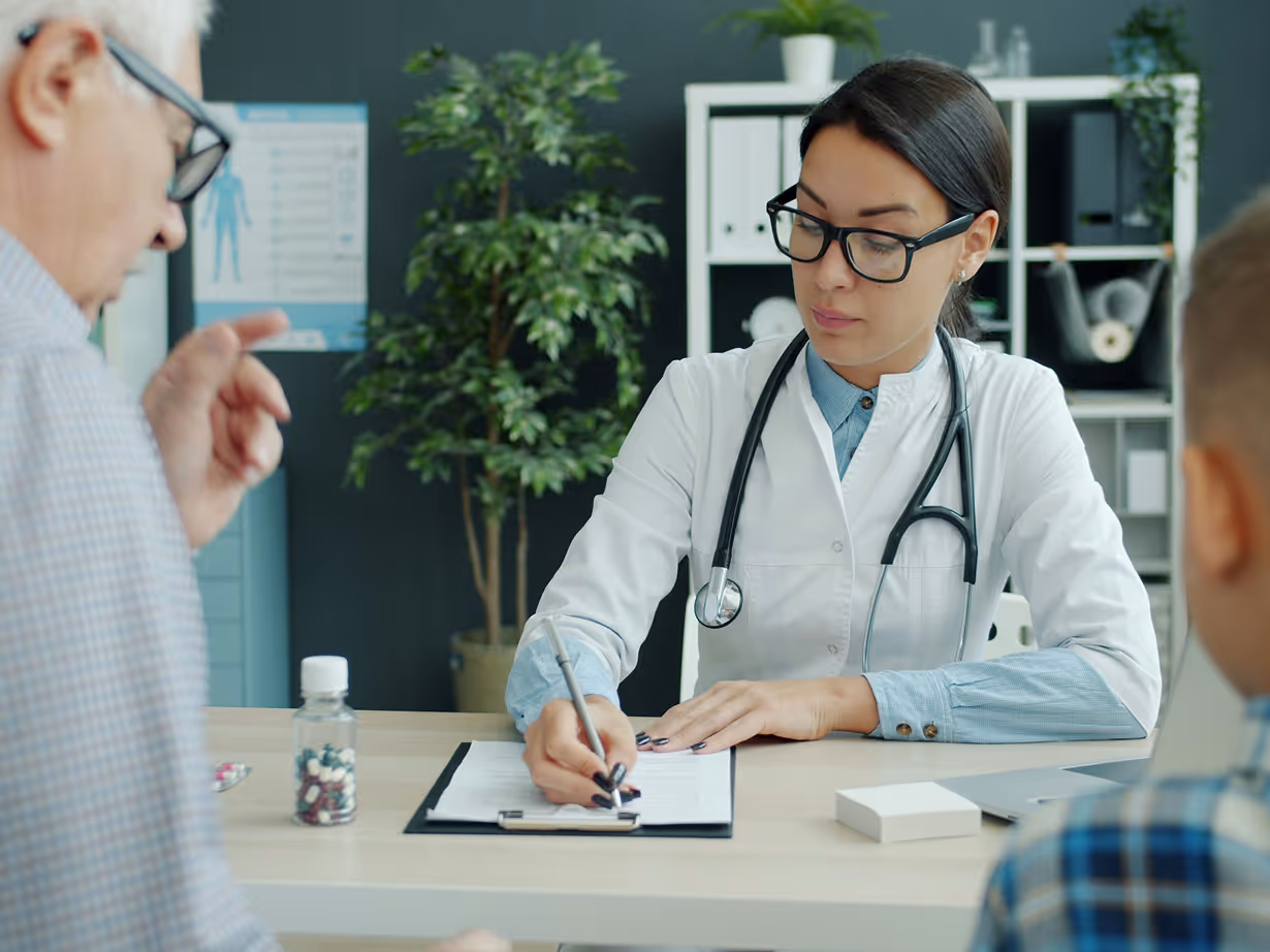 Female doctor wearing glasses and a stethoscope writing on a clipboard while consulting with an elderly man and a child in a medical office.