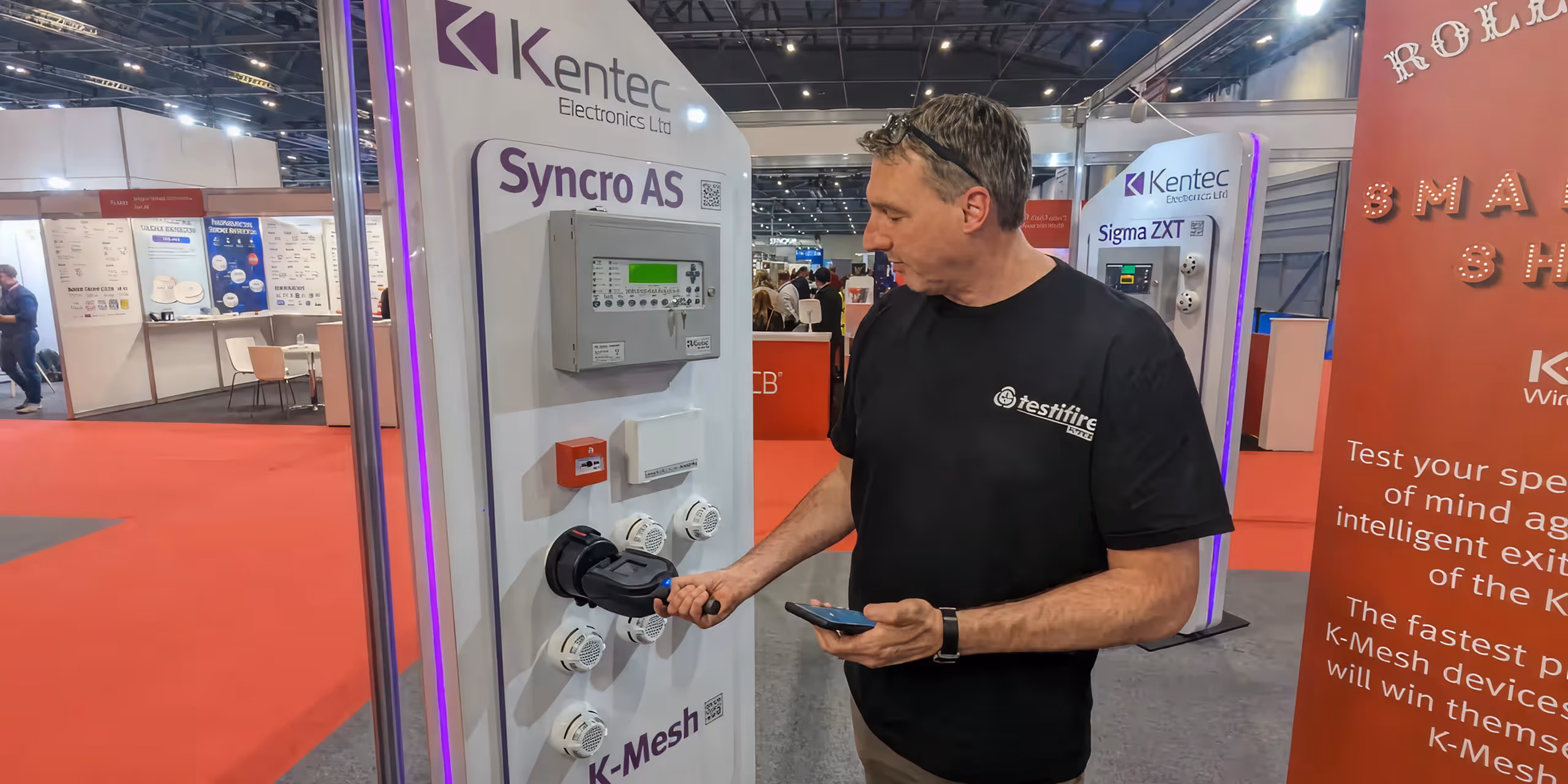 Man using a handheld device to test fire detection equipment at a Kentec Electronics Ltd exhibition booth.