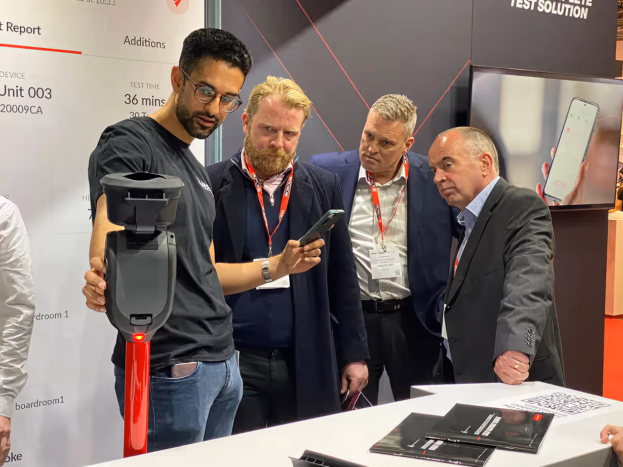 A man demonstrates a handheld device to three attentive men wearing conference badges at a trade show booth.