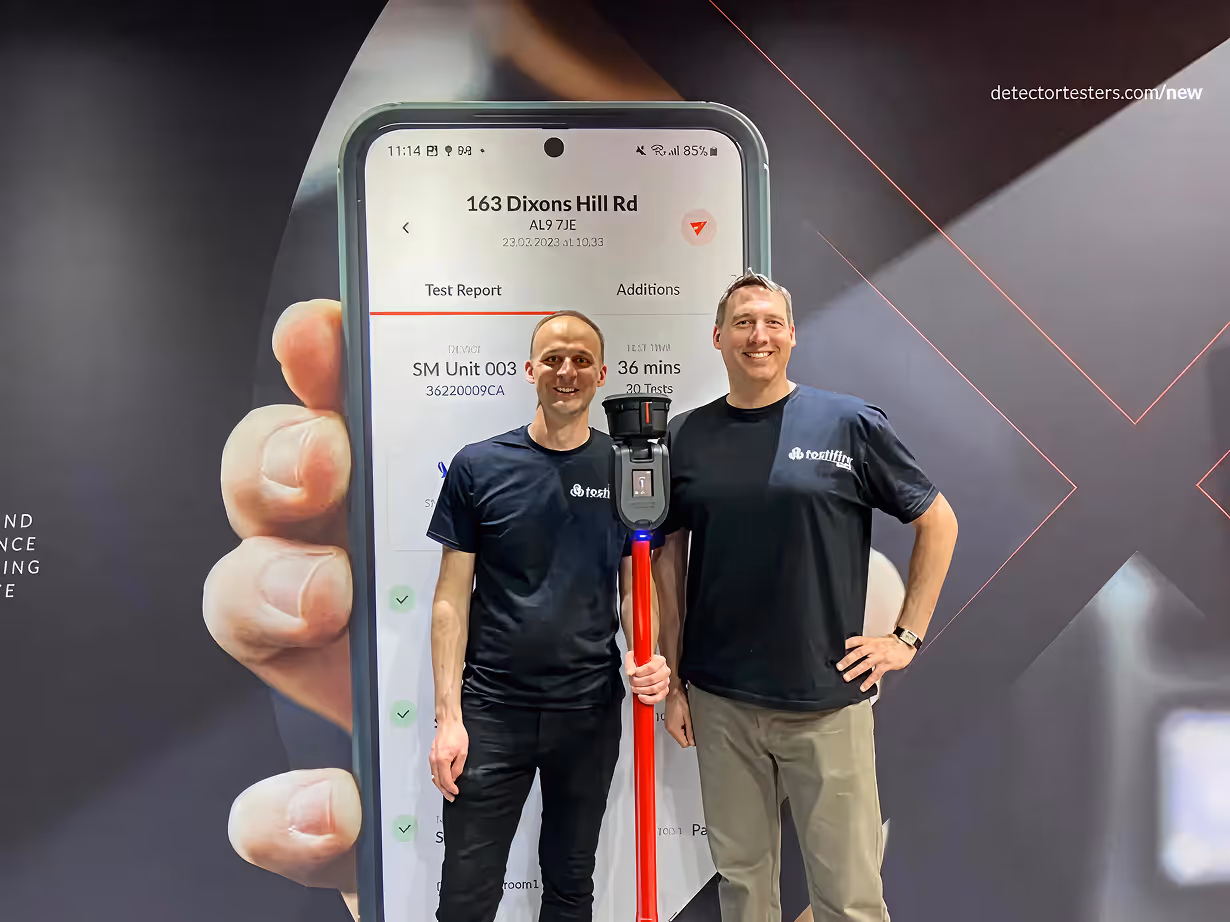 Two men wearing black Testifire t-shirts standing in front of a large smartphone display, one holding a red pole device with a black top.