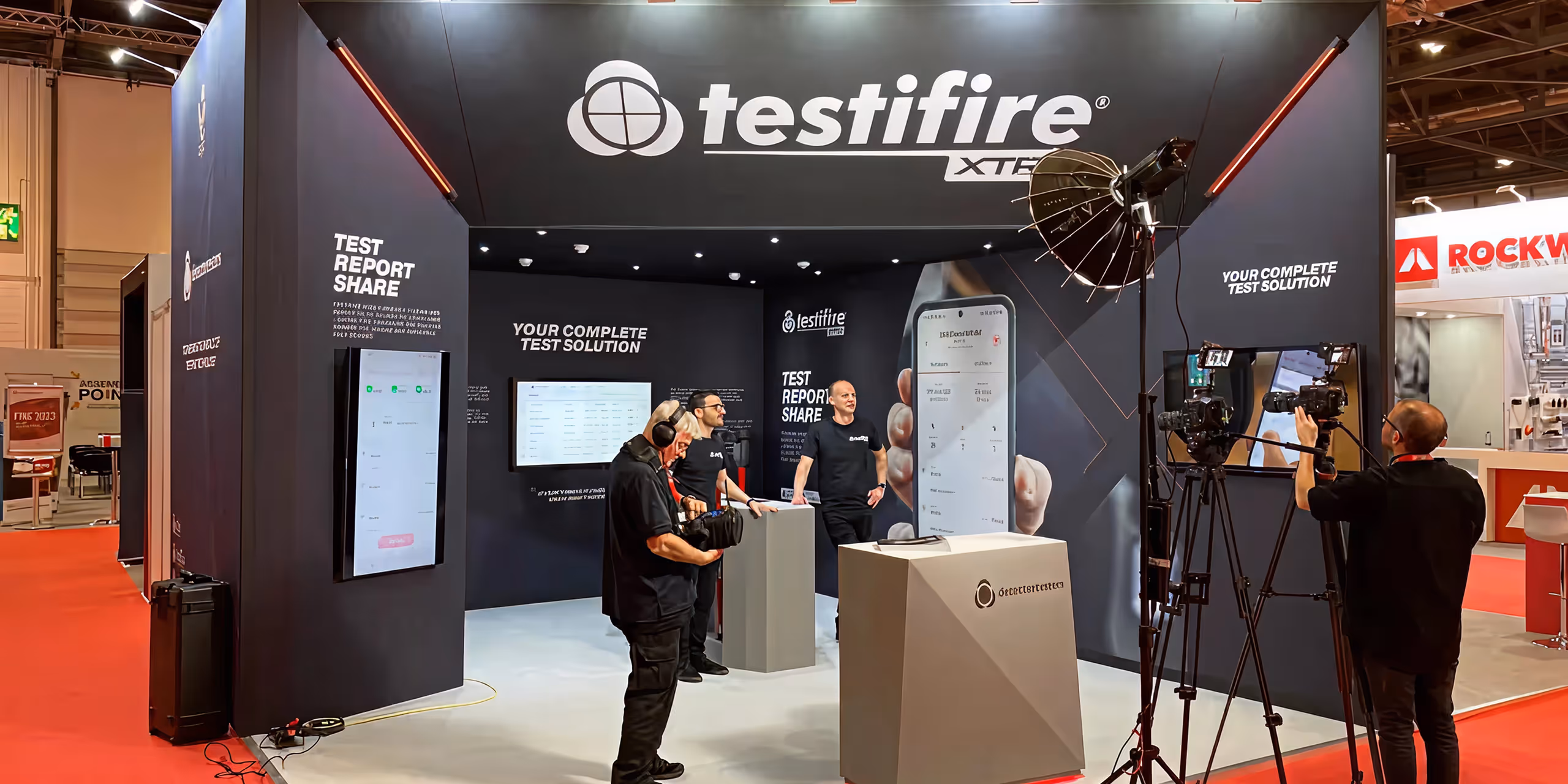Trade show booth for Testifire featuring three men in black shirts, cameras, studio lights, and digital screens displaying test report solutions.