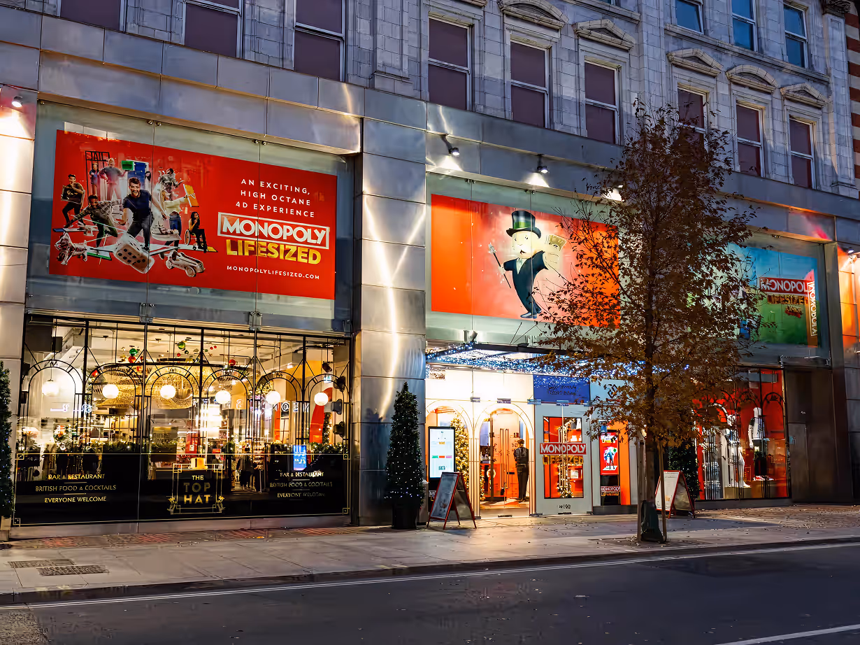 Exterior of a building with bright Monopoly Lifesized advertisements and a restaurant named The Top Hat with lit decor at night.