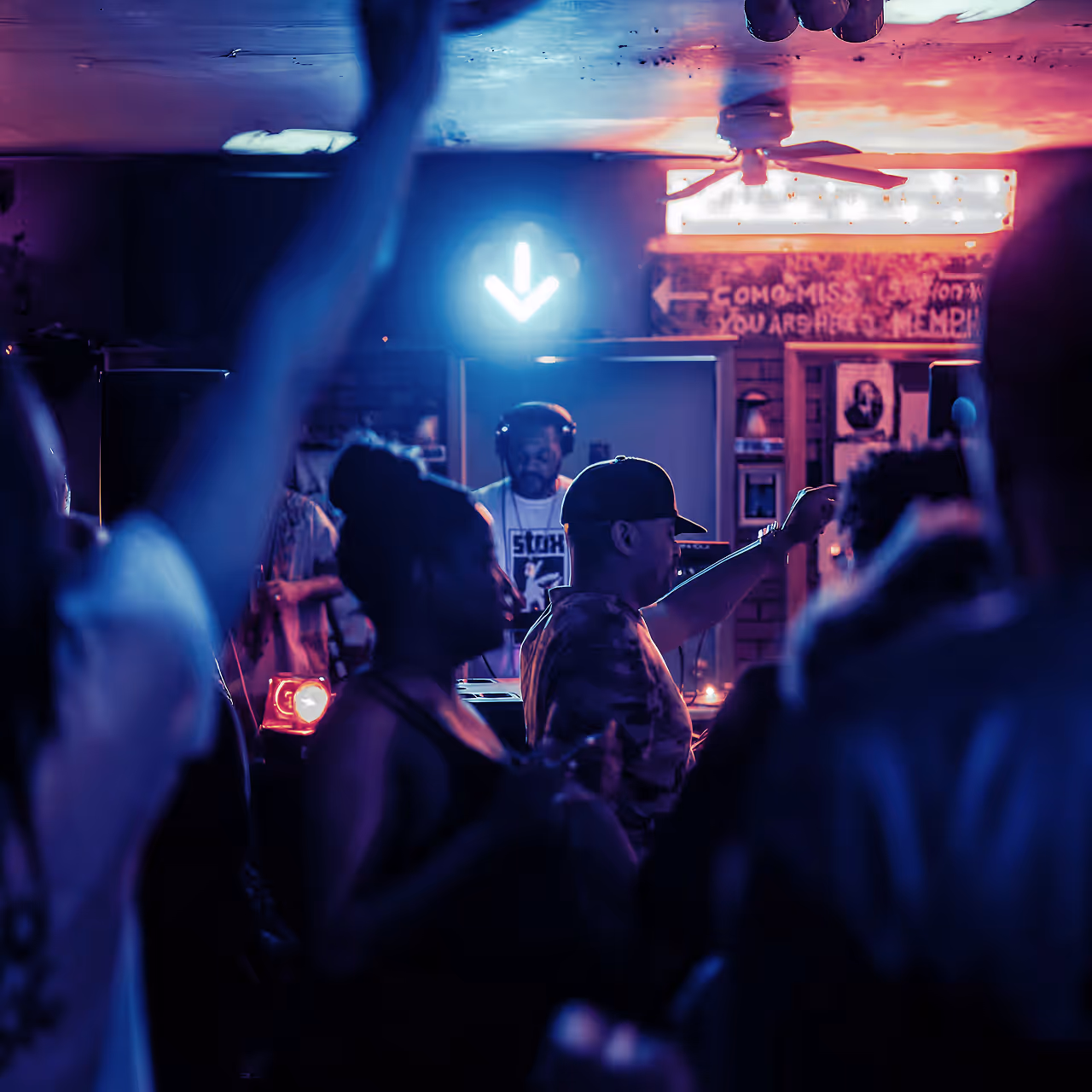 DJ wearing headphones playing music at a dimly lit nightclub with people dancing and raising their hands.