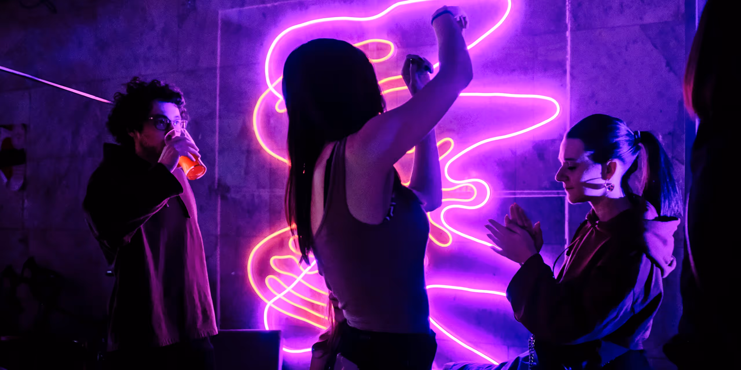 Three people in a dark room with purple neon light, one drinking from a cup, one dancing, and one clapping.