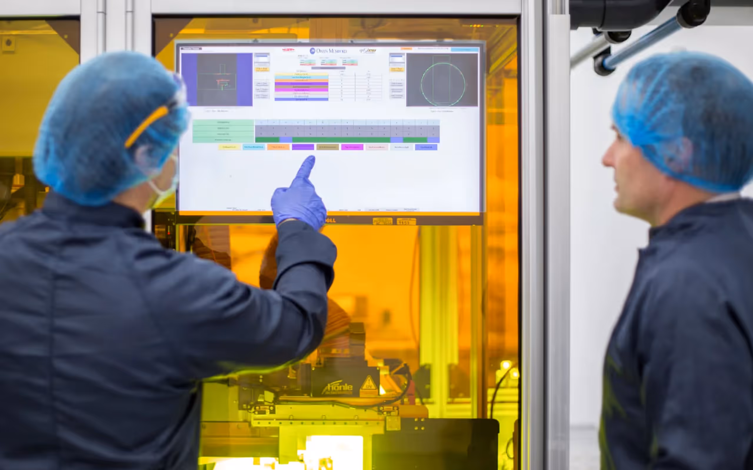 Two factory workers in blue hair nets and gloves operate and review data on a touchscreen monitor inside a laboratory or manufacturing environment.