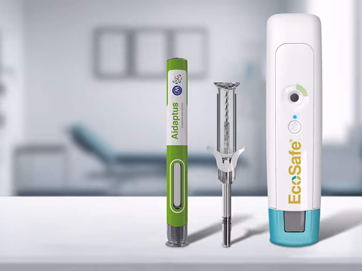 Three medical devices on a white surface with a blurred clinical setting in the background, including a green and white Aidaptus pen injector, a metal syringe, and a white and teal EcoSafe device.