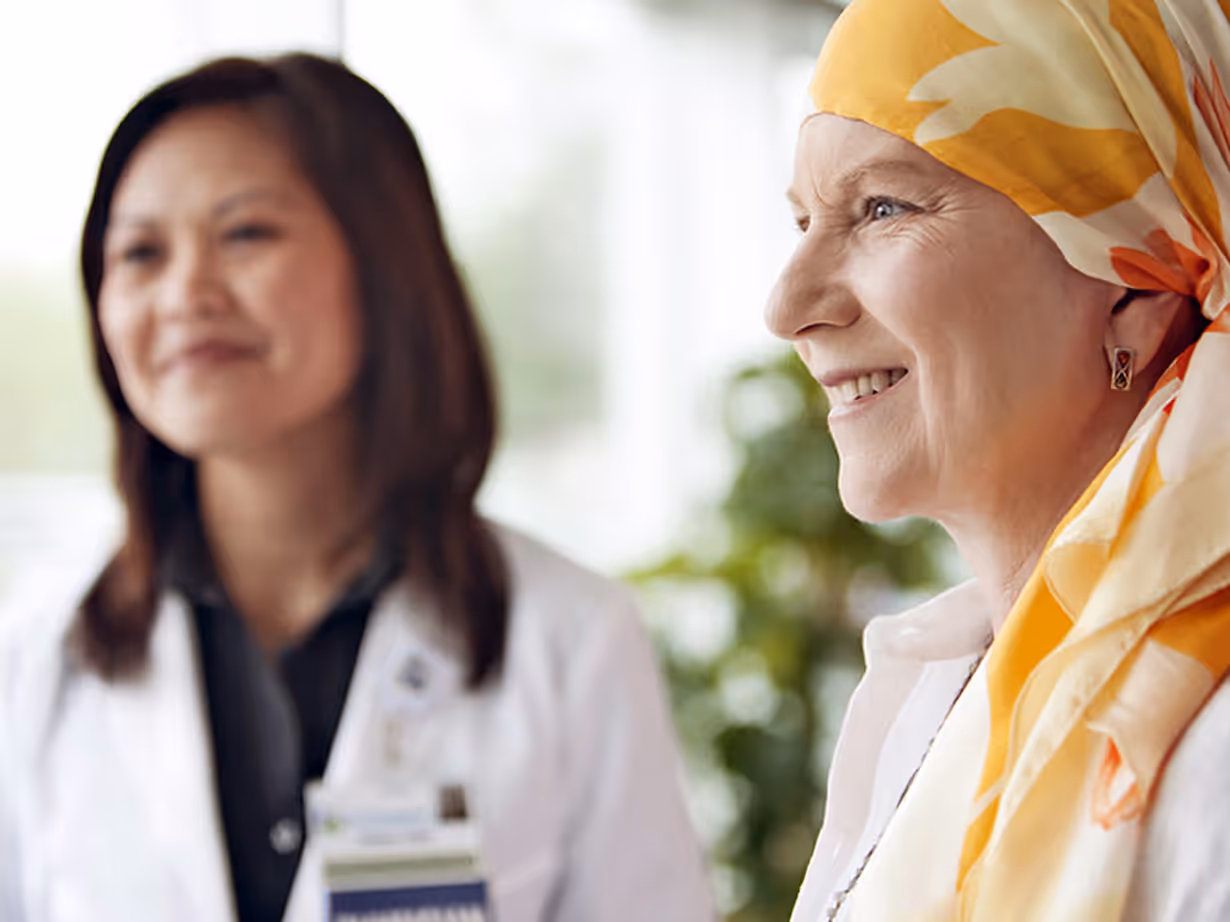 Smiling woman wearing a yellow and white headscarf, with a doctor in a white lab coat in the background.