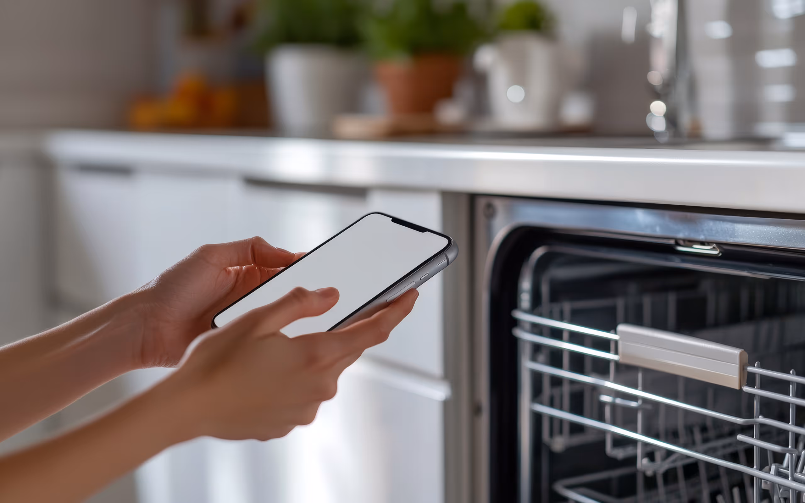 Person holding a smartphone with a blank screen in front of an open dishwasher in a modern kitchen.