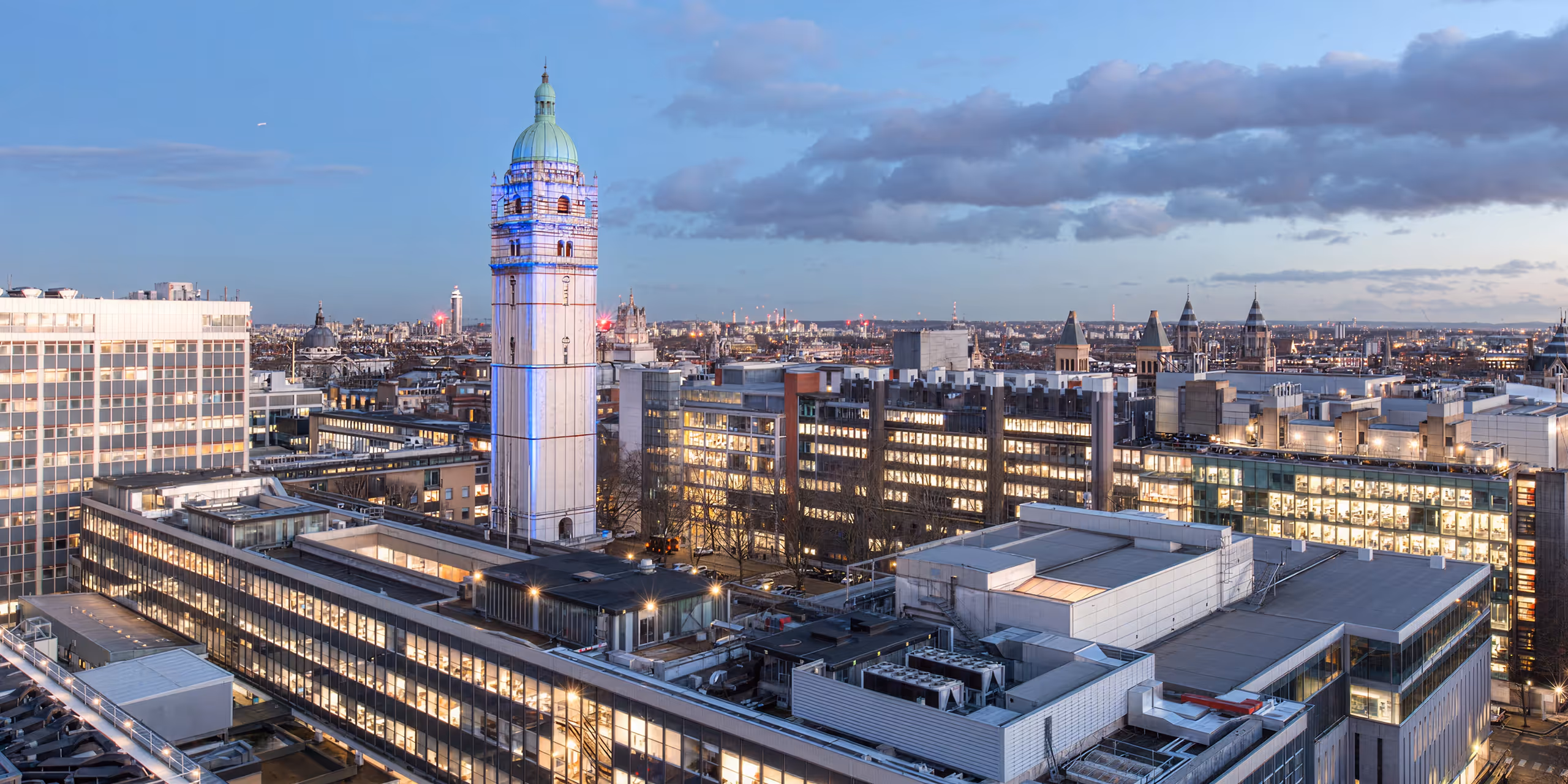 Skyline of London's urban area and Imperial College London at dusk featuring a tall white clock tower with a green dome, surrounded by modern office buildings with lit windows.