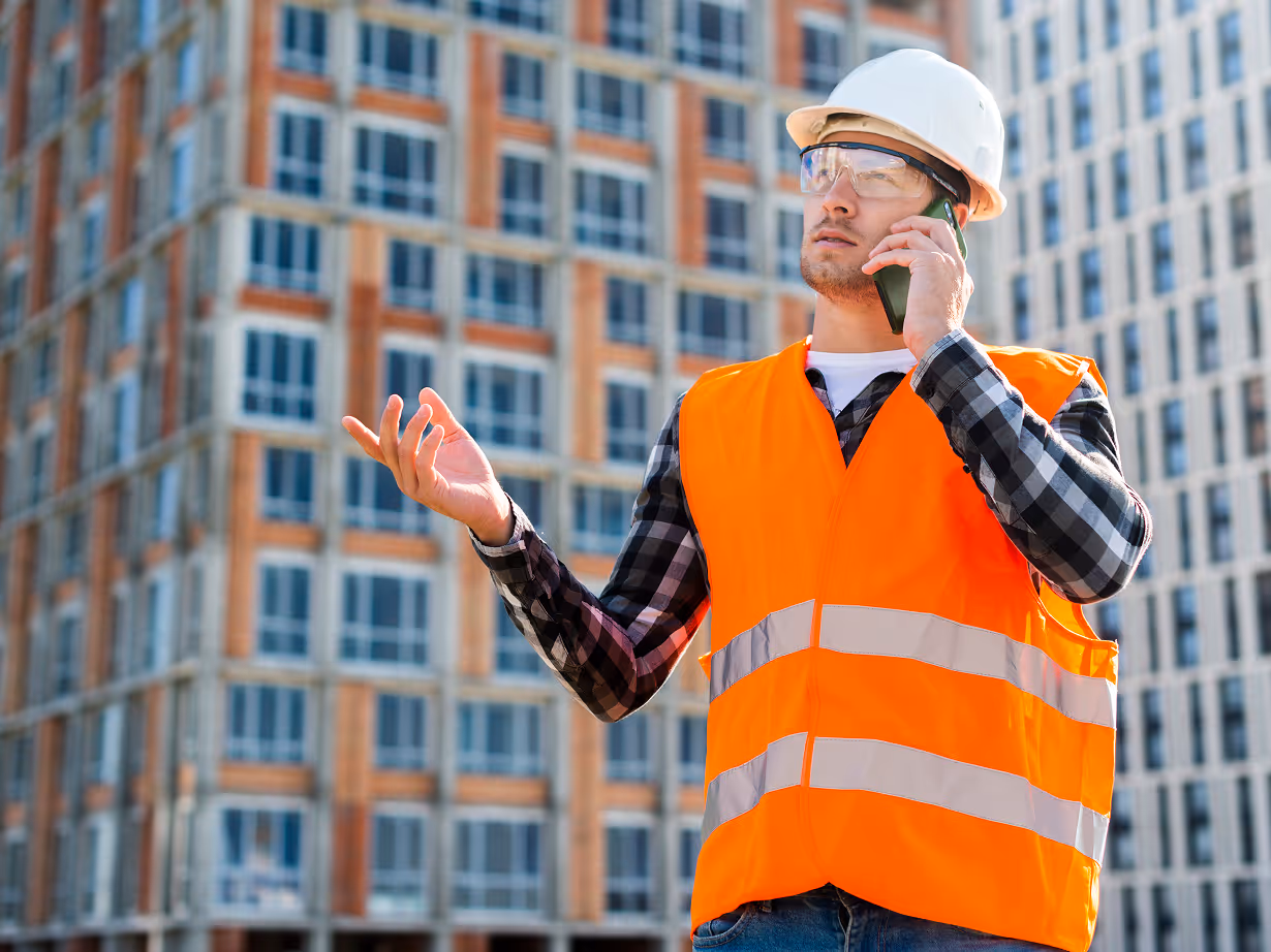 Construction worker wearing a white hard hat and orange safety vest talking on a phone in front of a high-rise building.