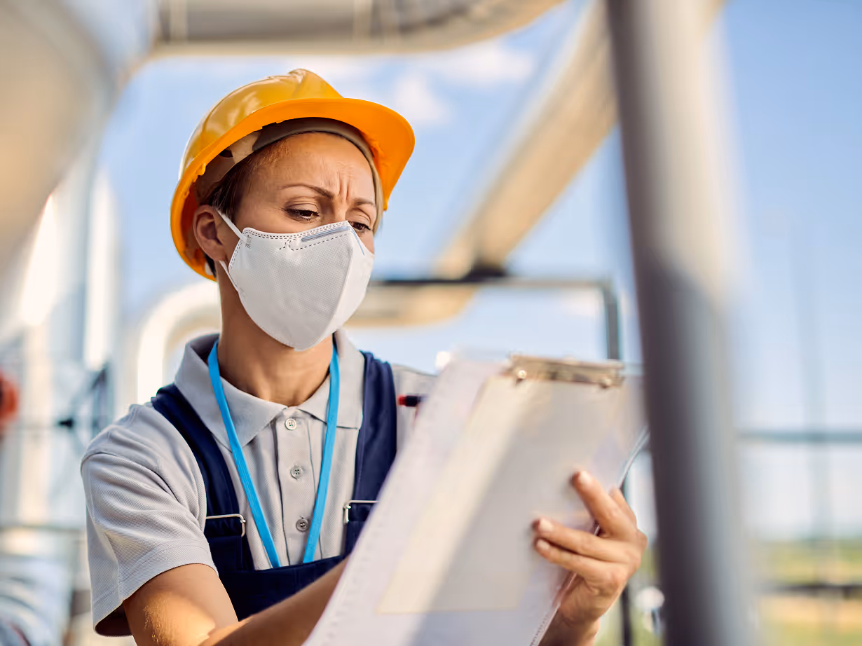 Woman in a yellow hard hat and face mask inspecting a clipboard at an industrial site.