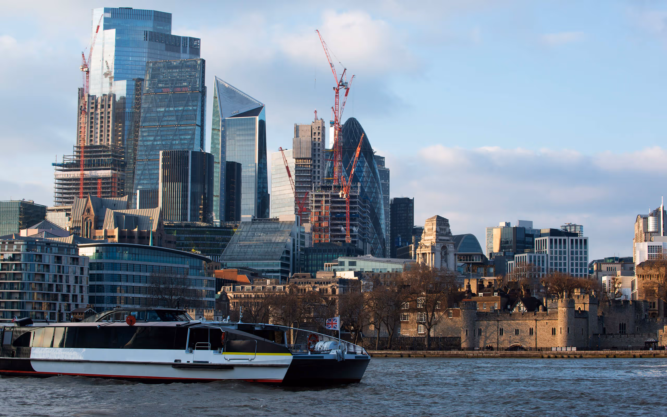 Cityscape of modern skyscrapers and historic buildings in London along a river with a black and white boat in the foreground.