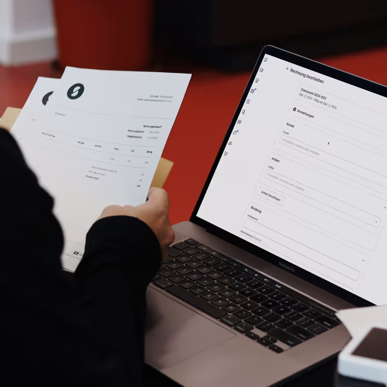 Person holding printed invoices while viewing an invoice editing page on a MacBook Pro laptop screen.