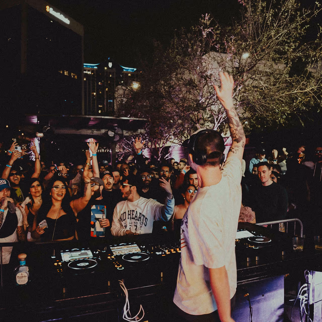 DJ wearing headphones and a white t-shirt plays music to an excited crowd at an outdoor nighttime event.