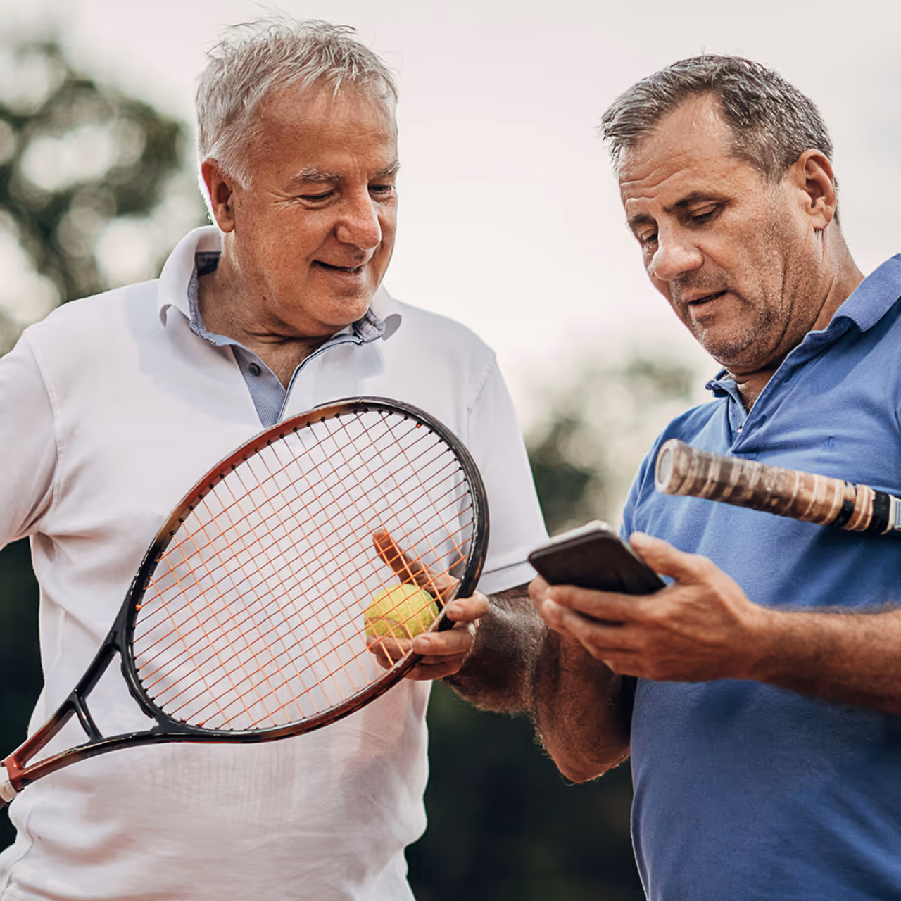 Two middle-aged men holding tennis rackets, one showing something on a smartphone to the other, outdoors.
